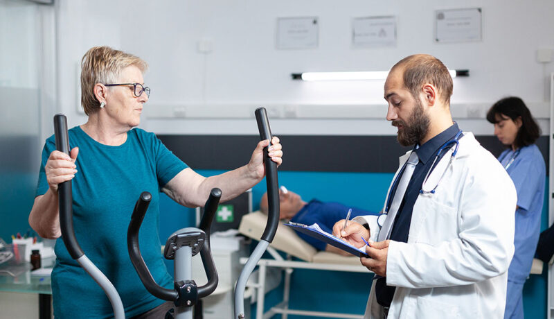 An Older Woman Using An Elliptical Machine While Talking With Her Physical Therapist Stages Of Cardiac Rehab