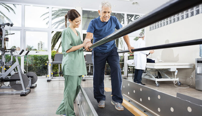 physical-therapy-for-fall-prevention A Senior Man Walking Between Parallel Bars Being Helped By A Physical Therapist During Physical Therapy For Fall Prevention