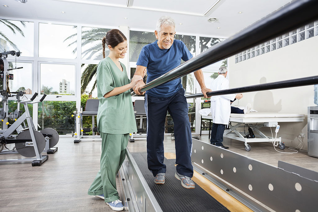 physical-therapy-for-fall-prevention A Senior Man Walking Between Parallel Bars Being Helped By A Physical Therapist During Physical Therapy For Fall Prevention